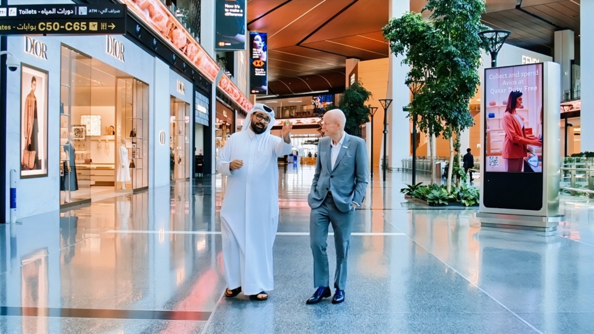 A high-resolution 4K image of two men walking through a modern shopping mall or airport terminal. One man wears traditional Middle Eastern attire, and the other is in a grey suit. They pass luxury stores like Dior, with directional signs overhead and a Qatar Duty Free advertisement displayed on a digital screen. The environment is clean, well-lit, and features potted plants.