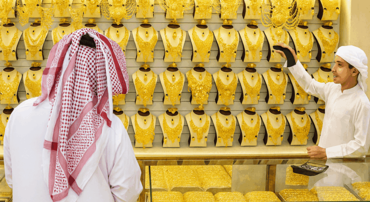 A jewelry shop filled with rows of gold necklaces and ornaments on display. A man wearing a white thobe and red-and-white checkered headscarf stands facing the display, while a shopkeeper in a white thobe and headscarf points at a necklace on the wall. The counter in front is also covered with trays of gold jewelry.