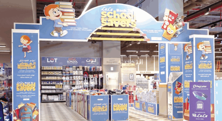 The image shows a LuLu Back to School Savers display in a store with various school supplies arranged on shelves and tables under a large blue arch. The arch features cartoon characters of children holding books and the text "LuLu Back to School Savers." Promotional signs in both English and Arabic highlight special offers and discounts for back-to-school items.