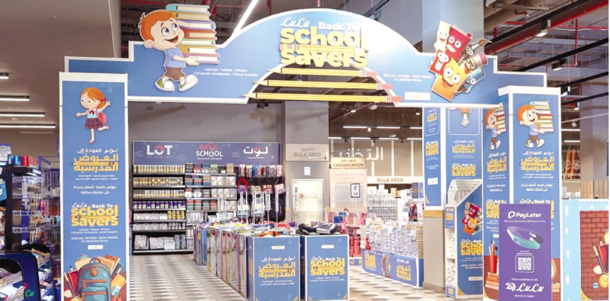 The image shows a LuLu Back to School Savers display in a store with various school supplies arranged on shelves and tables under a large blue arch. The arch features cartoon characters of children holding books and the text "LuLu Back to School Savers." Promotional signs in both English and Arabic highlight special offers and discounts for back-to-school items.